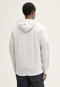 Felpa grigia chiara con cappuccio, dal finish texturizzato, tasca a marsupio e polsini a costine. Indossata sopra jeans scuri, vista da dietro.