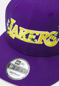 Gorra snapback morada con bordado en amarillo llamativo que dice "LAKERS," adornada con detalles en verde y una etiqueta circular de marca en el ala.