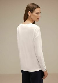 Donna con i capelli castani lisci indossa un top bianco a maniche lunghe con dettagli a bottoni sui polsini, rivolta di lato su uno sfondo beige.