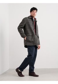 Braune kariert Jacke im Kragenstil mit zwei vorderen Taschen, getragen über einem kastanienbraunen Hemd und marineblauen Hosen. Braune elegante Schuhe runden das Outfit ab.