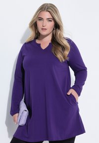 Fioletowa bluzka z długim rękawem i dekoltem w serek, wykonana z miękkiego materiału. Posiada luźny krój i boczne kieszenie. W połączeniu z pikowaną lawendową torebką.
