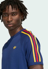 Mladić s kratkim dredovima, obučen u plavu majicu s žutim i crvenim prugama na ramenima, gleda ravno ispred sebe.