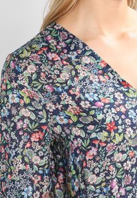 Kukkakuosiset paita tummansinisellä taustalla, jossa on monivärisiä kukkia ja vihreitä lehtiä. V-aukkoinen malli ja kevyen kankaan rakenne.