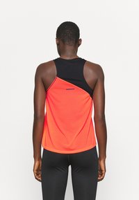 Person, der ein schwarzes ärmelloses Oberteil mit einem asymmetrischen orangen Überwurf auf dem Rücken trägt, kombiniert mit schwarzen Leggings, und von hinten schaut.