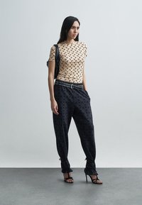 Tan oprijeta majica s abstraktnim črnim vzorcem, kombinirana z ohlapnimi črnimi hlačami z diskretnim tiskom logotipa in zvezanimi spodnjimi robovi. Črne sandale z naramnicami na peto.