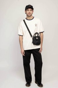 Tânăr îmbrăcat cu un tricou alb, pantaloni negri, sapcă neagră și geantă pe umăr neagră, stând în fața unui fundal deschis.