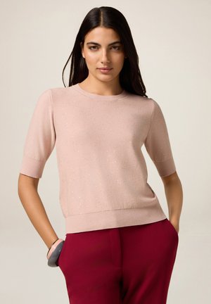 Vrouw met lang, donker haar in een lichtroze korte mouwen top met glitters en hooggetailleerde rode broek, handen in de zakken, neutrale achtergrond.
