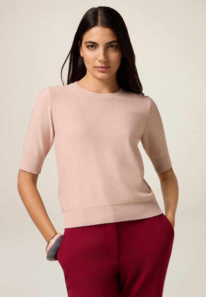 Kvinde med langt mørkt hår iført en lys pink t-shirt med korte ærmer og glimmer samt røde højtaljede bukser, hænderne i lommerne, neutral baggrund.