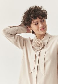 Blusa beige in tessuto liscio, con una grande rosa in tessuto al colletto e bordi legati, maniche lunghe e una vestibilità rilassata.