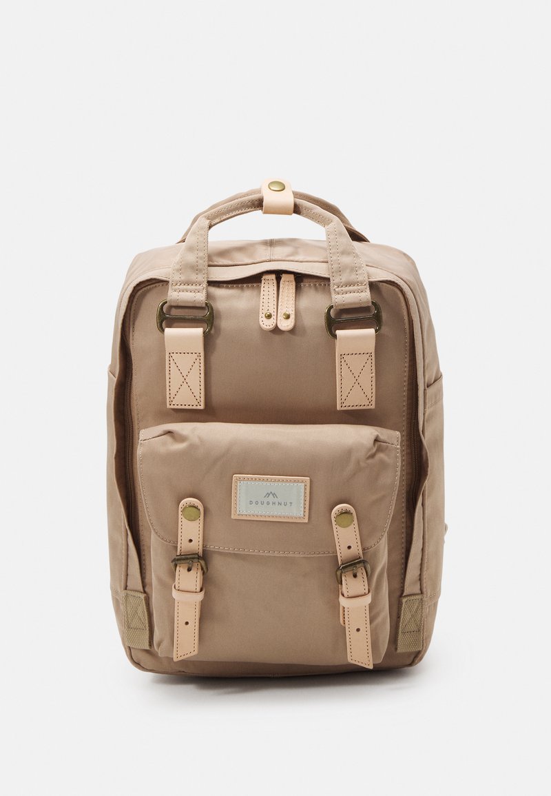 Tan fabric backpack with multiple pockets, top handle, and adjustable straps. Features a label and leather accents. Smooth texture.
