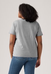 Person med krøllete mørkt hår iført en svart og hvitstripet kortermet skjorte og blå jeans, vendt bort fra kameraet.