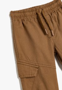 Pantalones cargo marrones con una cinturilla con cordón, bolsillo parche y tejido texturizado para mayor durabilidad. Las costuras en ángulo añaden un detalle de diseño.