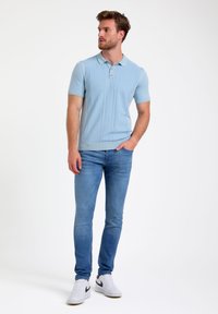 Camisa polo de punto azul claro con mangas cortas, cuello acanalado y detalles de costura vertical, combinada con jeans ajustados azul claro y zapatillas blancas.