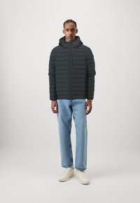 Sort pufferjakke med hætte, prydet med horisontale quiltede teksturer og en lynlåslomme på brystet, stylet med lyseblå jeans og hvide sneakers.