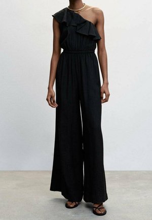 Femme portant une combinaison noire à une épaule avec un détail volanté, jambes larges et sandales noires, debout sur un sol gris clair.