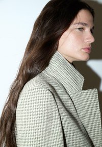 Svetlozeleni plašč z vzorcem houndstooth z visokim ovratnikom, ki ima strukturirano zasnovo in teksturirano blago, prikazan na nevtralni ozadju.