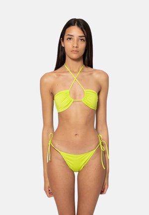 Donna con lunghi capelli scuri che indossa un bikini giallo a due pezzi con top allacciato al collo e slip con lacci laterali, in piedi su uno sfondo semplice.