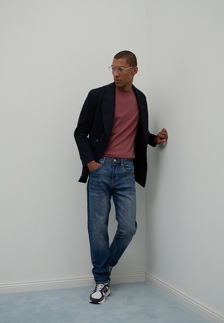 Uomo con occhiali, blazer blu scuro, camicia bordeaux, jeans blu e sneakers, che si appoggia in modo disinvolto a un muro bianco all'angolo.