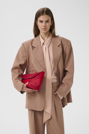 Γυναίκα με oversized μπεζ κοστούμι κρατώντας μια διπλωμένη κόκκινη τσάντα φάκελο πάνω σε απλό ανοιχτό φόντο.