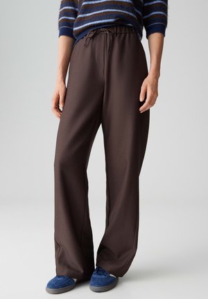 Personne portant un pantalon marron à jambes larges avec une taille à cordon, des baskets bleues et un pull rayé bleu, debout sur un fond clair.