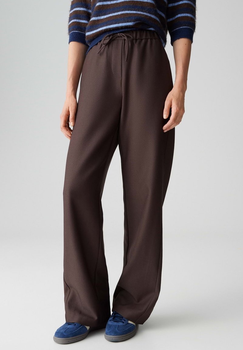 Personne portant un pantalon marron à jambes larges avec une taille à cordon, des baskets bleues et un pull rayé bleu, debout sur un fond clair.