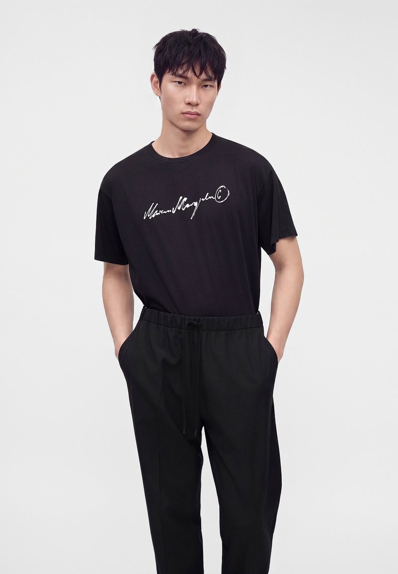 Jeune homme portant un pantalon noir et un T-shirt noir avec un texte blanc, debout devant un fond clair uni.