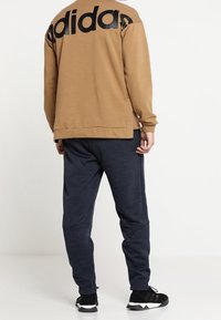 Sudadera marrón con gran logo negro de "adidas" en la parte posterior, combinada con joggers azul oscuro y zapatillas deportivas negras. Textura de tela suave.