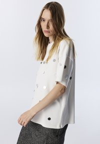 Weißes kurzärmeliges Blusenshirt mit silbernen, kreisförmigen Pailletten, die über die Vorderseite verstreut sind. Lockerer Schnitt und weicher Stoff.