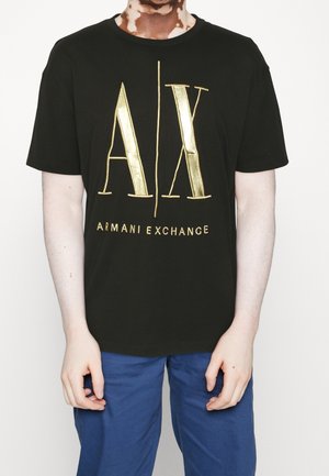 Osoba nosząca czarną koszulkę Armani Exchange z dużym złotym logo "A|X" i tekstem, w połączeniu z niebieskimi spodniami, stojąca na białym tle.