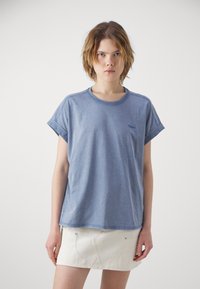 Tricou scurt cu maneci, de culoare albastru deschis, fabricat din bumbac moale, cu mansete rulote și guler rotund. Prezintă un mic logo în zona stângă a pieptului.