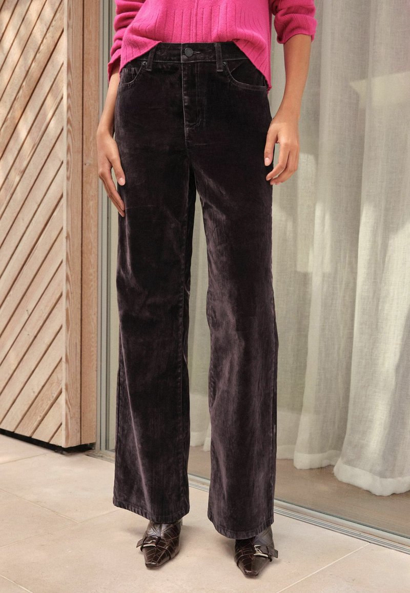 Melnas korduroja plata kāju bikses ar priekšējiem kabatām un pogu aizdari, papildinātas ar strukturētu rozā džemperi.