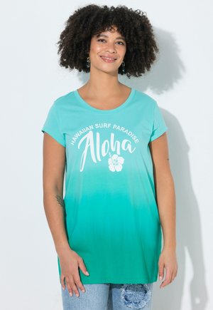 Frau mit lockigem Haar, die ein türkisfarbenes Ombre-T-Shirt mit der Aufschrift "Hawaiian Surf Paradise Aloha" und hellblaue Jeans mit Blumenstickerei trägt.
