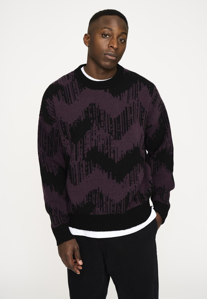 Strickpullover mit einem geometrischen schwarzen und lila Zickzackmuster, Rundhalsausschnitt, langen Ärmeln und einem strukturierten Stoff.