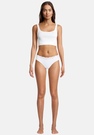 Frau steht barfuß da und trägt ein weißes, bauchfreies Tanktop sowie weiße Unterwäsche mit Spitzenbesatz vor einem einfarbigen Hintergrund.