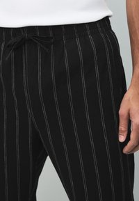 Schwarze Hose mit weißen vertikalen Streifen, ausgestattet mit einem elastischen Bund mit Kordelzug und Seitentaschen. Glatte Baumwollstofftextur.