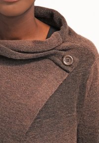 Brauner Strickpullover mit einem Cowl-Neck und einem strukturierten Muster, das mit einem braunen Knopf an der Schulter befestigt ist.