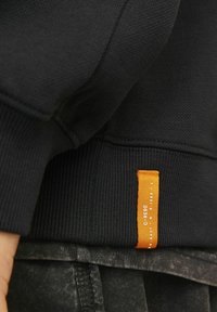Mustat teksturoidut hupparit, joissa on ribbaus hihansuissa, ja helmassa on ommeltu oranssi etiketti valkoisella tekstillä. Kangas sisältää geometrisia muotoja.