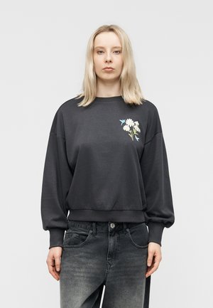 Donna bionda che indossa una felpa grigio scuro con ricami floreali e di uccelli sul petto a sinistra, abbinata a jeans neri slavati.