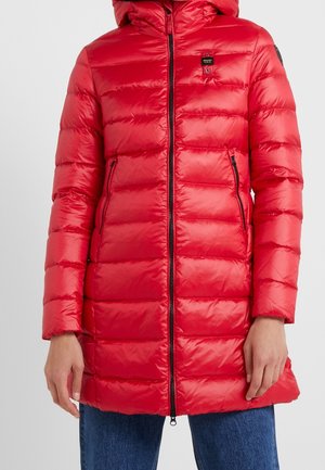 Manteau doudoune matelassé rouge avec capuche, fermeture éclair devant, poches latérales zippées, porté sur un jean bleu, visible du cou jusqu'au milieu des cuisses.