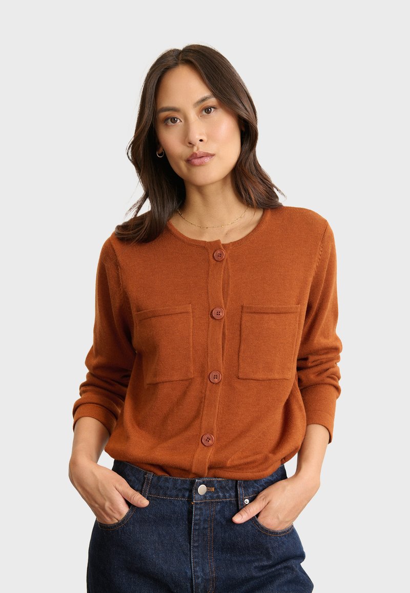 Cardigan color ruggine realizzato in morbido tessuto, con colletto rotondo, chiusura a bottoni e due tasche anteriori. Indossato con jeans di jeans scuri.