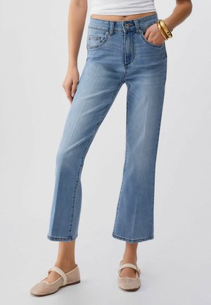 Frau trägt hellblaue, kurz geschnittene Jeans mit geradem Bein, weißes Oberteil, beigefarbene Mary Jane Schuhe aus Netzmaterial und goldene Armreifen an einem Handgelenk.