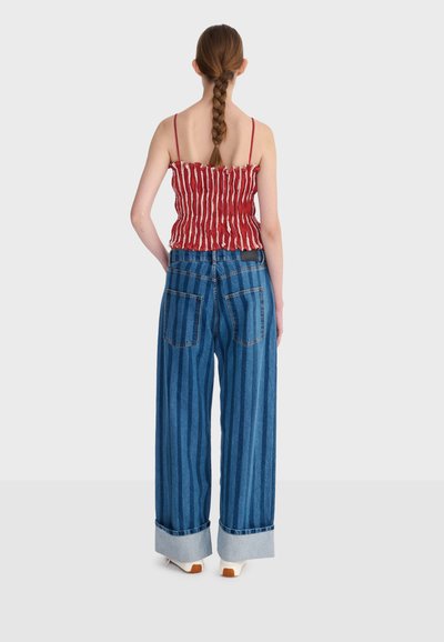 Femme aux cheveux tressés portant un haut sans manches rouge à rayures blanches et un jean à larges jambes bleu à rayures avec revers, debout de dos.