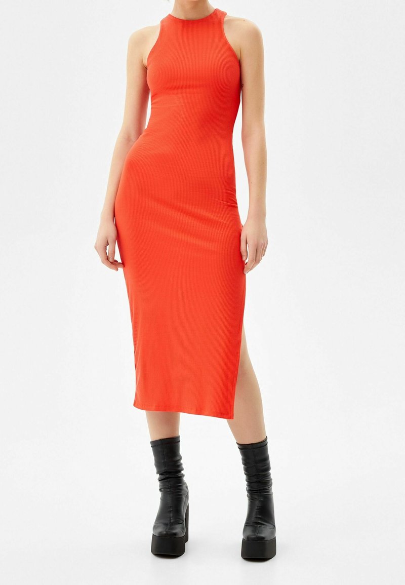 Femme portant une robe midi ajustée sans manches de couleur orange vif, associée à des bottines à plateforme noires, debout devant un fond blanc uni.