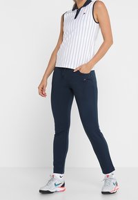 Ärmlös pikétröja i vitt med marinblå ränder och krage, kombinerad med marinblåa figurnära byxor och grå sneakers med färgade detaljer.