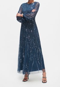 Lång marinblå klänning gjord av genomskinligt tyg med paljettbeklädda, virvlande mönster. Har långa, genomskinliga ärmar och en svävande siluett. Bärs med svarta klackar.