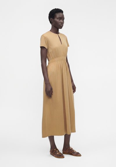 Femme debout portant une robe beige à manches courtes, longueur midi, avec une taille froncée et des sandales plates marron avec brides.