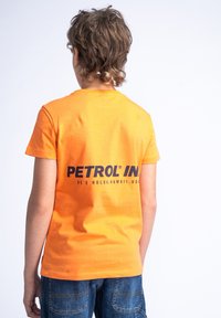 Camiseta de algodón naranja con mangas cortas, que presenta el texto negro "PETROL IN" y "HE'E HOLUA, HAWAI, EE. UU." impreso en la parte posterior.