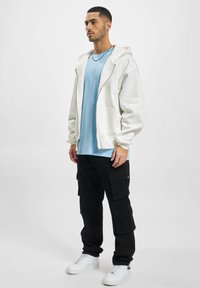 White zip-up hoodie over a light blue t-shirt, paired with black cargo pants and white sneakers. Simple design with functional pockets.