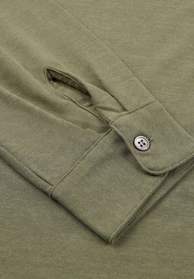 Polsino della camicia a maniche lunghe verde oliva con chiusura a bottone, caratterizzato da una texture morbida e un orlo arrotondato. Il materiale sembra leggero e liscio.