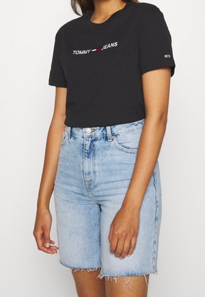 Kobieta w czarnej koszulce Tommy Jeans i jasnoniebieskich, wysokich szortach dżinsowych z postrzępionymi brzegami na jednolitym tle.